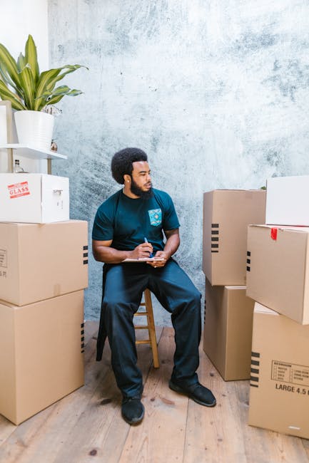 A man with dark curly hair and a beard is seated on a wooden stool inside a room with a light blue textured wall. He is wearing a dark blue t-shirt and matching trousers, and holding a clipboard with