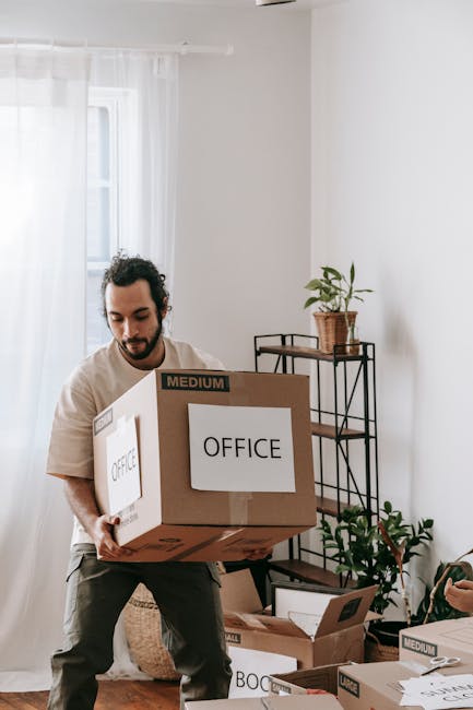 A man with dark curly hair and a beard is seated on a wooden stool inside a room with a light blue textured wall. He is wearing a dark blue t-shirt and matching trousers, and holding a clipboard with