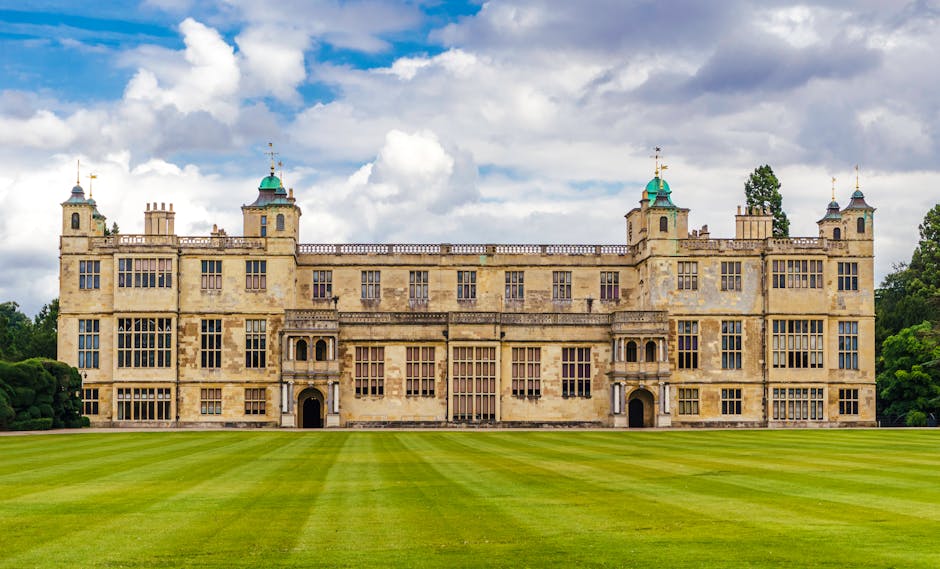 A large historic castle with multiple towers and turrets, constructed from light-colored stone, situated on a well-maintained grassy lawn with some trees on the right side. The building features numerous windows, decorative battlements, and a prominent central entrance with ornate detailing. The sky above is clear with a few clouds, and the scene is illuminated by natural daylight. This image captures the exterior of a grand, centuries-old estate, likely associated with heritage and historical significance, commonly involved in house removals or relocation services as part of a property transfer process. The setting suggests a peaceful environment suitable for moving or packing related to home relocation when viewed in the context of services offered by Man with Van Charlton.