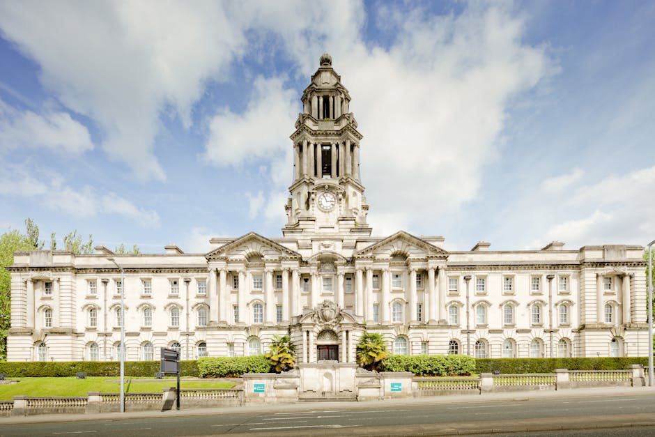 A large, historic white stone building with a central clock tower featuring multiple levels and ornate architectural details, situated behind a low hedge and accessible via paved sidewalk. The building's facade includes numerous evenly spaced windows, decorative columns, and intricate carvings near the entrance. In the foreground, a street with a curb and a few signs is visible, with the scene taken during daylight under partly cloudy skies. This exterior view of a prominent public or administrative building illustrates the setting where home relocation and furniture transport may require parking or access considerations, linked to the topic of whether a Greenwich Council permit is needed for van hire, as handled by Man with Van Charlton, a professional removals service provider.