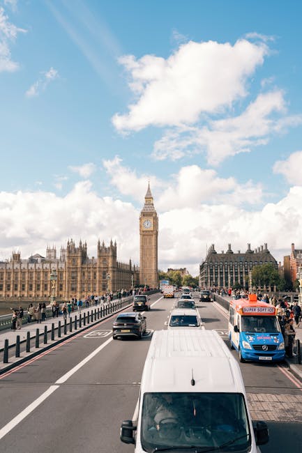 A busy street scene in central London featuring a view of the Houses of Parliament and the clock tower known as Big Ben under a partly cloudy sky. Several vehicles, including a white van, are parked or moving along the road, with some cars visible in the distance. Pedestrians are walking on the pavement beside the street, and a blue 'Super Soft Ice' delivery truck is parked on the right side. The image captures the urban environment during daylight hours, with traffic and foot traffic typical of a city centre. This scene relates to house removals and logistical planning involved in home relocation services, such as those provided by Man with Van Charlton, as they navigate the busy streets of Greenwich for loadings or deliveries.