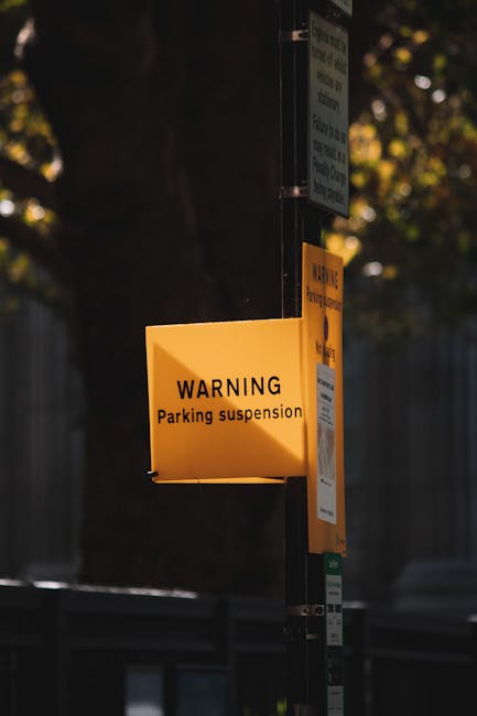 A close-up view of a bright yellow warning sign attached to a black metal pole, displaying the message 'WARNING Parking suspension' in black text on the yellow background. The sign is part of a temporary parking restriction notice related to house removals or moving services in Charlton, as indicated by the context of the webpage. Behind the pole, there are additional signs, including a pale blue and green informational sign, and the background features blurred trees and a fence, suggesting an outdoor urban environment. The lighting suggests late afternoon or early evening, with shadows cast on the sign, highlighting the importance of parking permissions during home relocation activities carried out by Man with Van Charlton, supporting efficient furniture transport and packing and moving logistics.