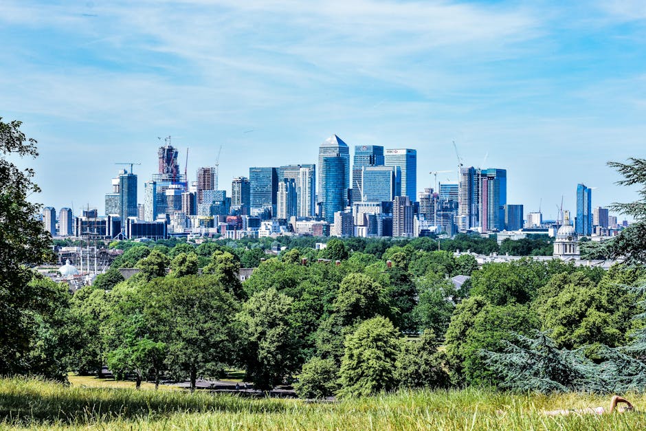 A panoramic view of a modern city skyline featuring numerous high-rise buildings with glass facades, some under construction with cranes visible, set against a bright blue sky with scattered clouds. In the foreground, a lush green park area with dense trees and well-maintained grass provides a natural contrast to the urban environment. The scene appears to be taken from a higher vantage point within a residential area, possibly during daytime, capturing the contrast between home surroundings and city development. This image relates to house removals and moving services, highlighting the importance of careful planning when relocating furniture and belongings from a home within or near such urban landscapes. Man with Van Charlton, a professional removals company, offers expertise in managing the logistics of house moves, including navigating parking restrictions like those implemented by Greenwich Council, for a smooth home relocation experience.