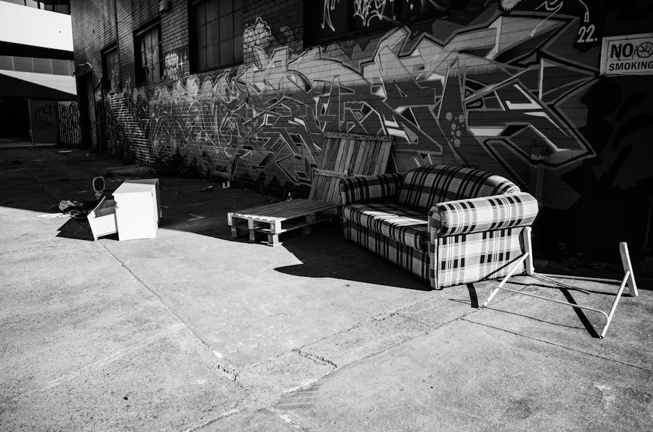 A black and white photograph showing an outdoor loading area adjacent to a building with a graffiti-covered wall. In the foreground, there are disassembled furniture items including a patterned fabric