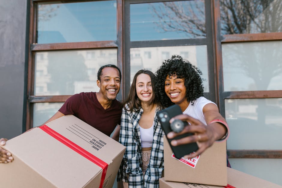Three individuals, two women and one man, stand outdoors in front of a building with large glass windows and wooden frames during daylight. They are smiling and holding unpacked cardboard boxes, some with packing labels and plastic straps, indicating a home relocation process. One woman with curly hair is taking a selfie with a smartphone, while the others hold boxes, one of which appears to contain household items or clothing. The scene captures a moment of packing and moving preparations, with natural daylight illuminating the scene, reflecting typical activities associated with furniture transport and packing during a house move. The individuals are casually dressed, and the background shows a partially blurred urban environment. This image is suitable for illustrating moving services provided by [COMPANY_NAME], such as packing, loading, and logistics involved in house removals, as highlighted on the page titled '[PAGE_TITLE]' at [PAGE_URL].