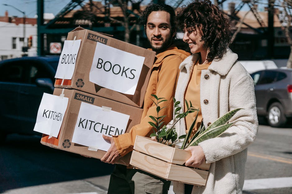 A man and woman are walking outdoors in a parking lot, each carrying stacks of moving boxes and plants. The man is holding two cardboard boxes labeled 'BOOKS' and 'KITCHEN,' which are stacked one on top of the other, and appears to be in the process of a home relocation. The woman is holding a wooden crate containing potted plants, likely prepared for transport as part of packing and moving activities. Behind them, a blue car is visible along with parked vehicles and a building with an outdoor structure, suggesting a loading area for a house move. The scene shows clear daylight, with the individuals dressed in casual outdoor clothing appropriate for the moving process. The boxes are secured with tape, and some labels are printed on paper attached to the boxes, indicating their contents. The environment sustains a professional yet approachable scene consistent with house removal and furniture transport services provided by Man with Van Charlton, supporting efficient packing and loading processes during a scheduled move.