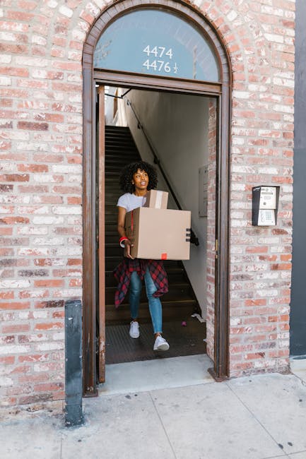 Three individuals, two women and one man, stand outdoors in front of a building with large glass windows and wooden frames during daylight. They are smiling and holding unpacked cardboard boxes, some with packing labels and plastic straps, indicating a home relocation process. One woman with curly hair is taking a selfie with a smartphone, while the others hold boxes, one of which appears to contain household items or clothing. The scene captures a moment of packing and moving preparations, with natural daylight illuminating the scene, reflecting typical activities associated with furniture transport and packing during a house move. The individuals are casually dressed, and the background shows a partially blurred urban environment. This image is suitable for illustrating moving services provided by [COMPANY_NAME], such as packing, loading, and logistics involved in house removals, as highlighted on the page titled '[PAGE_TITLE]' at [PAGE_URL].