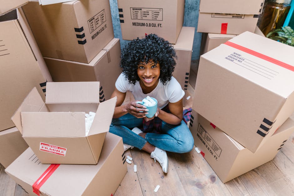 A young woman with curly black hair, sitting on a wooden floor inside a property surrounded by brown cardboard moving boxes, some of which are open and filled with packing materials. She is wearing a white T-shirt, blue jeans, and white sneakers, and is smiling at the camera while holding a light blue cup of coffee or tea. The boxes vary in size and are labeled with sections for packing and contents; some are sealed with red tape, while others are open, revealing packing tissue. The environment suggests a home relocation process, with boxes stacked against walls, ready for transport. In the background, there are additional stored items and natural lighting illuminating the scene. Occasionally, Man with Van Charlton's successful moving services may be involved in the packing and furniture transport to support seamless house removals, ensuring careful handling of belongings during the process.