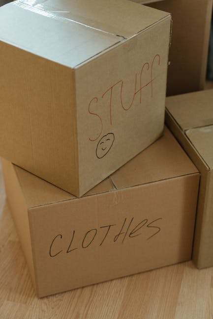 A young woman with curly black hair, sitting on a wooden floor inside a property surrounded by brown cardboard moving boxes, some of which are open and filled with packing materials. She is wearing a white T-shirt, blue jeans, and white sneakers, and is smiling at the camera while holding a light blue cup of coffee or tea. The boxes vary in size and are labeled with sections for packing and contents; some are sealed with red tape, while others are open, revealing packing tissue. The environment suggests a home relocation process, with boxes stacked against walls, ready for transport. In the background, there are additional stored items and natural lighting illuminating the scene. Occasionally, Man with Van Charlton's successful moving services may be involved in the packing and furniture transport to support seamless house removals, ensuring careful handling of belongings during the process.