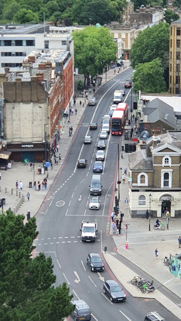 An aerial view of Woolwich Road in Charlton showing a street with multiple parked and moving vehicles, including cars and a red double-decker bus, on a two-lane road with a slight curve. On the left side, there are pedestrians walking along the pavement and storefronts, while on the right, a small white building with a pitched roof is situated next to bicycles parked near a bicycle rack. The street is lined with tall, leafy green trees providing shade, and residential and commercial buildings are visible in the background. The scene captures typical urban traffic and foot traffic during daylight hours, with an emphasis on the road environment where home relocation and furniture transport services by Man with Van Charlton might operate during house removals or moving logistics.