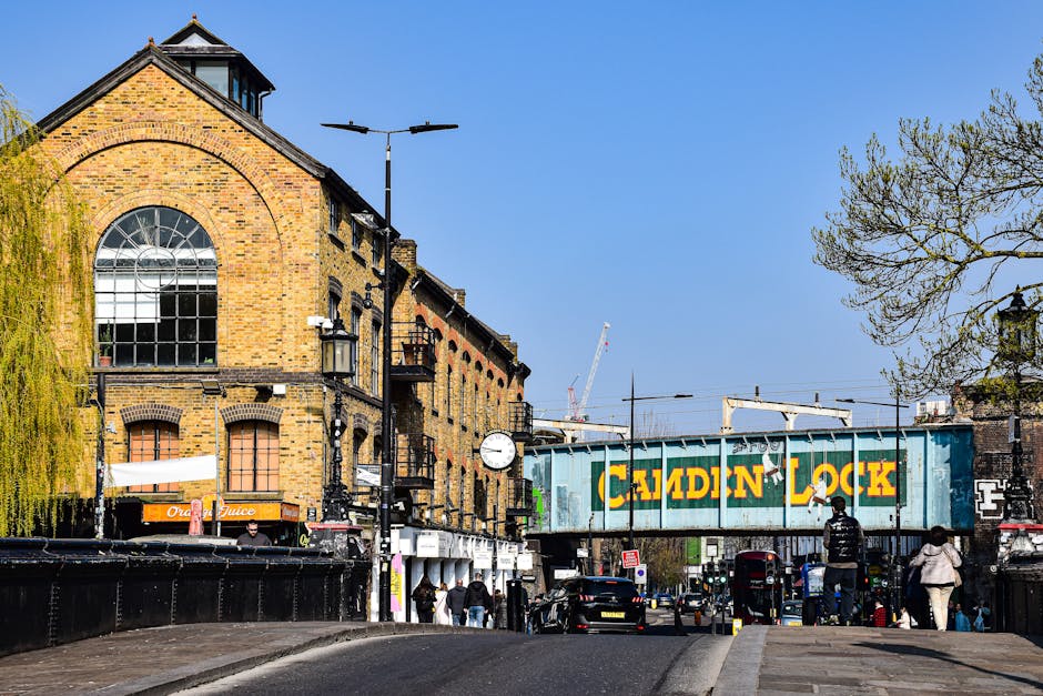 A busy street scene in Woolwich, featuring a row of brick buildings with large arched windows on the upper floors. On the pavement, several people are gathered nearby, some walking and others standing. A blue freight container with the words 'Calden Lock' painted in yellow is mounted on an old railway bridge structure that runs across the street, indicating a railway or freight line nearby. In the foreground, a black van is parked, likely used for furniture transport during home relocation, with its rear door partially open. A crane and construction equipment are visible in the background, suggesting ongoing development or moving activities in the area. The street is lined with trees, some with budding leaves, and the sky above is clear and bright, highlighting the urban setting where packing, moving, and logistics take place, supported by companies like Man with Van Charlton specializing in removals.