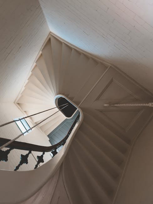 An indoor spiral staircase with wooden steps and black decorative balustrades leads down to a lower floor in a residential property. The staircase is enclosed by white-painted walls with a textured finish. Sunlight filters through a small window on the landing, illuminating the area. At the top of the staircase, there is a small landing space with a closed white door and a section of wall visible. The image captures the stairs during the process of home relocation handled by Man with Van Charlton, demonstrating the careful navigation of household furniture and packing materials through narrow spaces as part of the loading process for furniture transport and removal services.