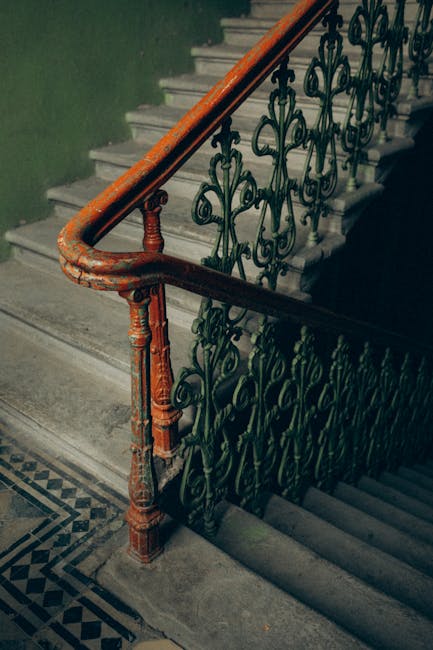 A close-up view of a vintage indoor staircase in a Victorian-style property, displaying a worn wooden handrail painted orange and an ornate black wrought iron balustrade with intricate scrollwork. The staircase has concrete steps and the surrounding wall is painted green. The patterned tiled floor at the base of the staircase indicates a traditional interior. This scene captures the staircase during a home relocation process, where furniture or boxes from the upper floors may be transported carefully. Man with Van Charlton, a professional removals company, is experienced in handling such historic staircases safely during packing, loading, and furniture transport, ensuring minimal damage during the loading process in residential properties like these.