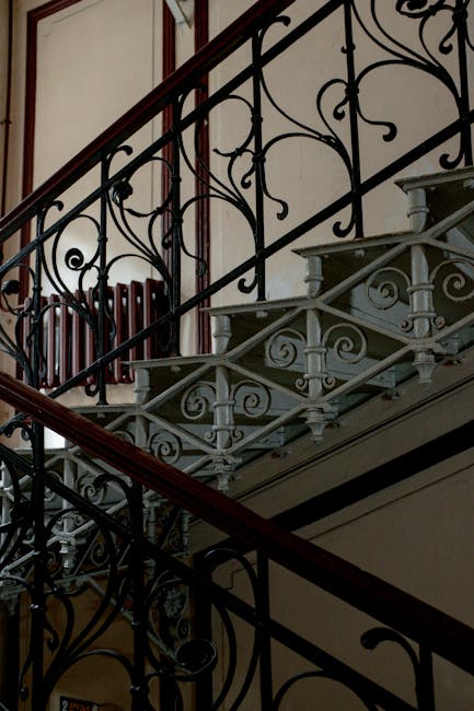 An indoor spiral staircase with wooden steps and black decorative balustrades leads down to a lower floor in a residential property. The staircase is enclosed by white-painted walls with a textured finish. Sunlight filters through a small window on the landing, illuminating the area. At the top of the staircase, there is a small landing space with a closed white door and a section of wall visible. The image captures the stairs during the process of home relocation handled by Man with Van Charlton, demonstrating the careful navigation of household furniture and packing materials through narrow spaces as part of the loading process for furniture transport and removal services.