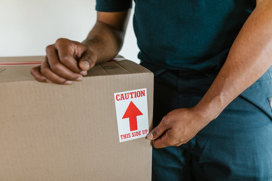 A person wearing a dark green T-shirt and dark trousers is seen inside a property, guiding a large cardboard box with a red and white 'CAUTION THIS SIDE UP' label attached. The individual is holding the box steady, with one hand gripping the top edge and the other supporting the side. The cardboard box is positioned near a doorway, suggesting it is being prepared for transport as part of a home relocation or furniture transport process. The box is sealed, and the label indicates careful handling during the packing and moving operation. Behind the person, a glimpse of the interior environment is visible, with natural lighting illuminating the scene. The image reflects the loading process typically handled by services like Man with Van Charlton, focusing on proper packing and safe lifting to ensure furniture and belongings are securely moved from one location to another.