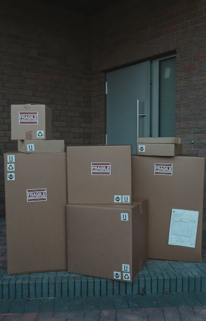 A person wearing a dark green T-shirt and dark trousers is seen inside a property, guiding a large cardboard box with a red and white 'CAUTION THIS SIDE UP' label attached. The individual is holding the box steady, with one hand gripping the top edge and the other supporting the side. The cardboard box is positioned near a doorway, suggesting it is being prepared for transport as part of a home relocation or furniture transport process. The box is sealed, and the label indicates careful handling during the packing and moving operation. Behind the person, a glimpse of the interior environment is visible, with natural lighting illuminating the scene. The image reflects the loading process typically handled by services like Man with Van Charlton, focusing on proper packing and safe lifting to ensure furniture and belongings are securely moved from one location to another.