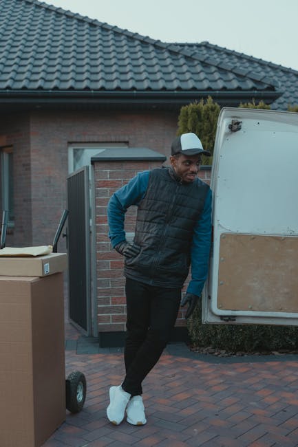 A man dressed in a blue long-sleeved shirt, black padded vest, and black trousers is standing outdoors on a paved driveway beside a white van with beige paneling, which is parked near a brick house wi